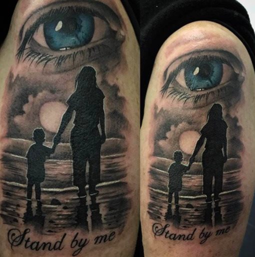 Tatuajes para Madres Hijos y Familia realista artistico en brazo ojo celeste madre e hijo mirando un atardecer en el mar con la frase Stand by me Quedate a mi lado