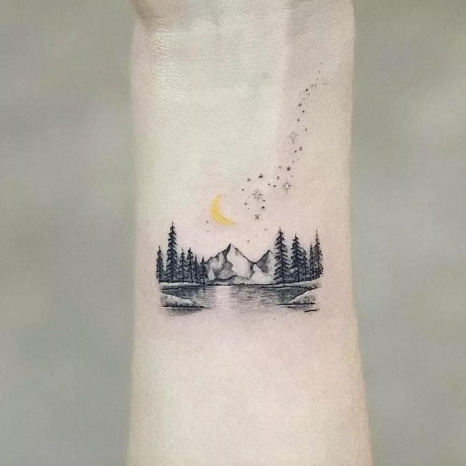 Forearm tattoo of alpine scene with pine trees and yellow crescent moon