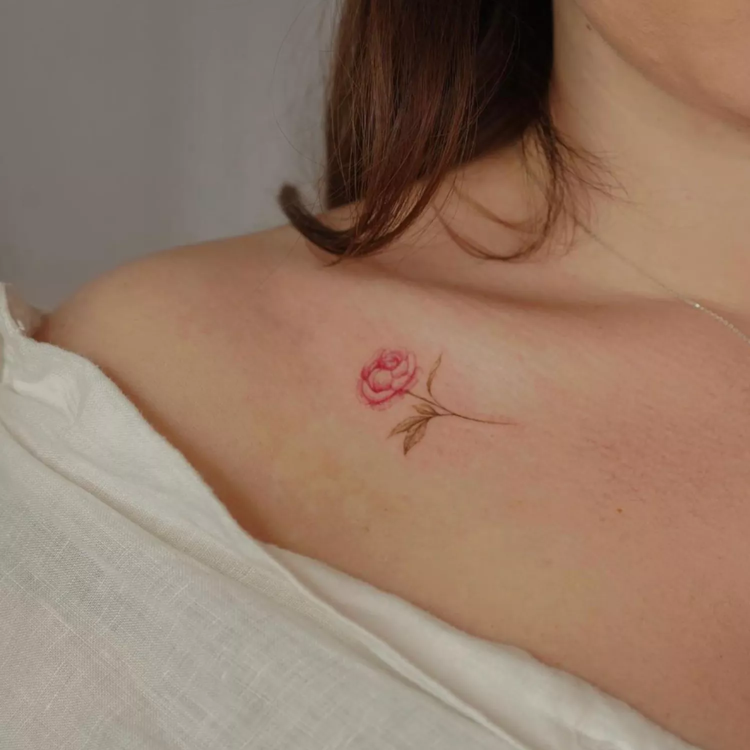 zoomed-in image of model with peony tattoo on collarbone with green stem and leaves and pink/red petals